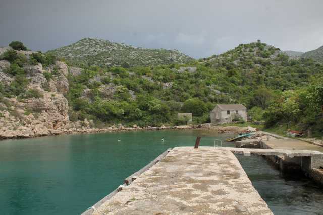 Sanfte Hügel umgeben einen kleinen, ruhigen Hafen, dessen Wasser in einem tiefen, klaren Türkis schimmert. Am linken Rand erhebt sich eine steinerne Klippe, die mit üppiger, grüner Vegetation durchzogen ist. Die unterschiedlichen Grüntöne von Laub und Sträuchern schaffen eine harmonische Kulisse entlang der Felsen, deren raue Textur durch die unterschiedlichen Lichtverhältnisse stark hervorgehoben wird. Am rechten Bildrand steht ein schlichtes, graues Gebäude, dessen Dach leicht geneigt ist. Die Oberfläche des Hauses scheint von der Zeit gezeichnet und trägt Spuren von Verwitterung. Neben dem Gebäude befindet sich ein kleiner, bewaldeter Bereich, der einen Kontrast zu den kargen Felsen bildet. Auf der rechten Seite sind einige Boote sichtbar, die am Steg liegen und darauf warten, ins Wasser geschoben zu werden. Der Weg zum Wasser führt über eine breite, graue Steinmauer, die sich in die Bucht hineinzieht. Auf diesem Weg sind einige unregelmäßige Steine zu sehen, die die Struktur unterbrechen und eine natürliche, rustikale Ästhetik erzeugen. Ein rostiger Pfahl ragt am Ende des Stegs in die Höhe, was den Eindruck von einem langen Zeitabschnitt vermittelt, in dem hier Aktivitäten stattfanden. Im Hintergrund sind die sanften Hügel sichtbar, die mit dichtem Grün bewachsen sind und im oberen Bereich flach auslaufen. Der Himmel ist von grauen Wolken bedeckt, die eine gewisse Dramatik vermitteln und die Lichtverhältnisse im Bild beeinflussen. Das Gesamtbild strahlt eine ruhige und unberührte Schönheit aus, die den Betrachter in die pazifistische Stimmung des Ortes hineinzieht.