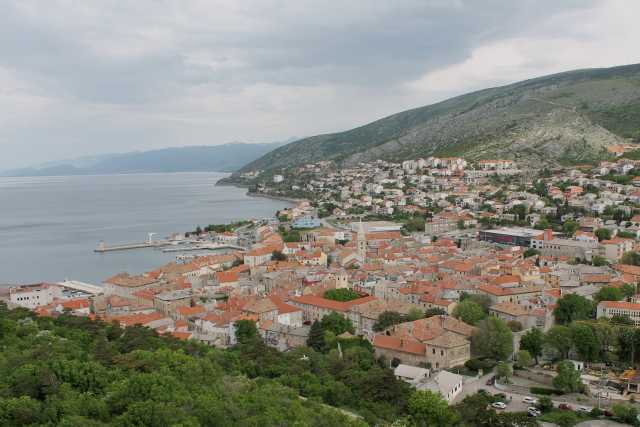 Das Bild zeigt eine malerische Küstenstadt, eingebettet zwischen einem grünen Hügel und dem Meer, unter einem bewölkten Himmel. Das Bild zeigt eine atemberaubende Aussicht auf eine Küstenstadt, die sich an den Hängen eines Hügels entlang erstreckt und auf das Meer blickt. Im Vordergrund ist eine üppige, grüne Vegetation zu sehen, die die Stadt von der Aussichtsplattform trennt. Die Stadt selbst ist ein dichtes Gefüge aus niedrigen, hauptsächlich ockerfarbenen und rötlich-braunen Häusern mit roten Ziegeldächern. Die Gebäude sind eng beieinander gebaut und zeigen eine Vielzahl von Größen und Stilen, was auf eine lange Geschichte und organische Entwicklung hinweist. Einige Gebäude wirken moderner, andere älter und traditioneller. Eine Kirchturmspitze ist prominent im Zentrum der Stadt zu sehen. Im unteren mittleren Bereich des Bildes verläuft eine Straße, die sich durch das Stadtzentrum schlängelt und den Zugang zu den verschiedenen Teilen der Stadt ermöglicht. Am unteren Ende des Bildes ist ein kleiner Hafen mit einigen Booten sichtbar. Das Meer ist ruhig und dunkelgrau, und der Himmel ist bedeckt mit dicken, grauen Wolken, die eine leicht düstere, aber dennoch ruhige Atmosphäre schaffen. Im Hintergrund erheben sich Berge, die teilweise im Nebel liegen. Die Gesamtkomposition des Bildes suggeriert ein Gefühl von Ruhe und Geschichte, mit einem Kontrast zwischen der natürlichen Schönheit und der bebauten Umgebung. Die Farben sind gedämpft, was zur allgemeinen Stimmung beiträgt.
