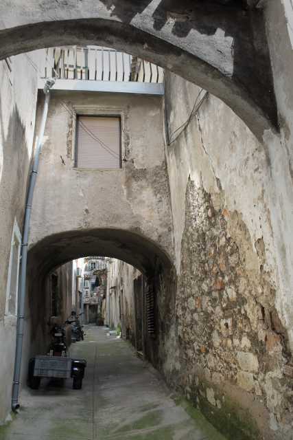 Hier ist eine einfache Beschreibung des Bildes: Das Bild zeigt eine enge Gasse in einer alten Stadt. Ein Steinbogen überspannt die Gasse und gibt den Blick auf den weiteren Verlauf frei. Die Wände sind alt und verwittert, mit abblätterndem Putz und freiliegendem Stein. Am linken Rand der Gasse steht ein kleiner Wagen. Insgesamt vermittelt das Bild einen Eindruck von Alter, Geschichte und dem Charme einer kleinen, verwinkelten Gasse.