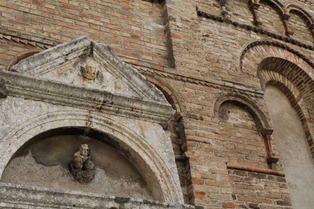 Hier ist eine einfache Beschreibung des Bildinhalts: Das Bild zeigt einen Ausschnitt eines alten, steinernen Gebäudes. Im Vordergrund ist eine steinerne Fassade mit einem Bogen und einem dreieckigen Giebel darüber zu sehen. An der Wand des Gebäudes befinden sich mehrere dekorative Elemente und architektonische Details wie weitere Bögen. Die Struktur besteht aus roten Backsteinen und Steinen, was ihr ein rustikales und historisches Aussehen verleiht. Das Bild ist aus einer schrägen Perspektive aufgenommen, die Details der Architektur hervorhebt.