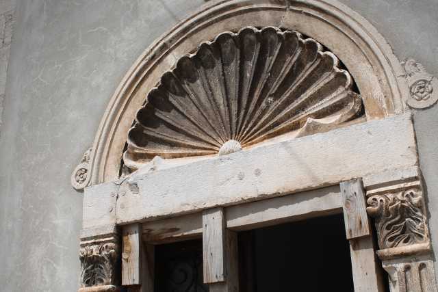 Dieses Bild zeigt einen verwitterten Steintürrahmen mit einem muschelförmigen Ornament über der Tür. Der Rahmen ist aus hellem Stein gefertigt und zeigt Anzeichen von Alterung und Verfall. Das Muschelmotiv ist detailliert gearbeitet und deutet auf einen möglichen Bezug zur Meereswelt oder zur klassischen Architektur hin. Die Tür selbst ist mit Holzbrettern provisorisch repariert, was auf einen Zustand des Verfalls oder der Renovierung hinweist. Die Kombination aus verziertem Stein und provisorischer Holzreparatur erzeugt einen interessanten Kontrast zwischen alter Pracht und aktuellem Zustand, und unterstreicht die Geschichte des Bauwerks. Das Bild wirkt insgesamt ruhig und evoziert ein Gefühl von Geschichte und Vergänglichkeit.