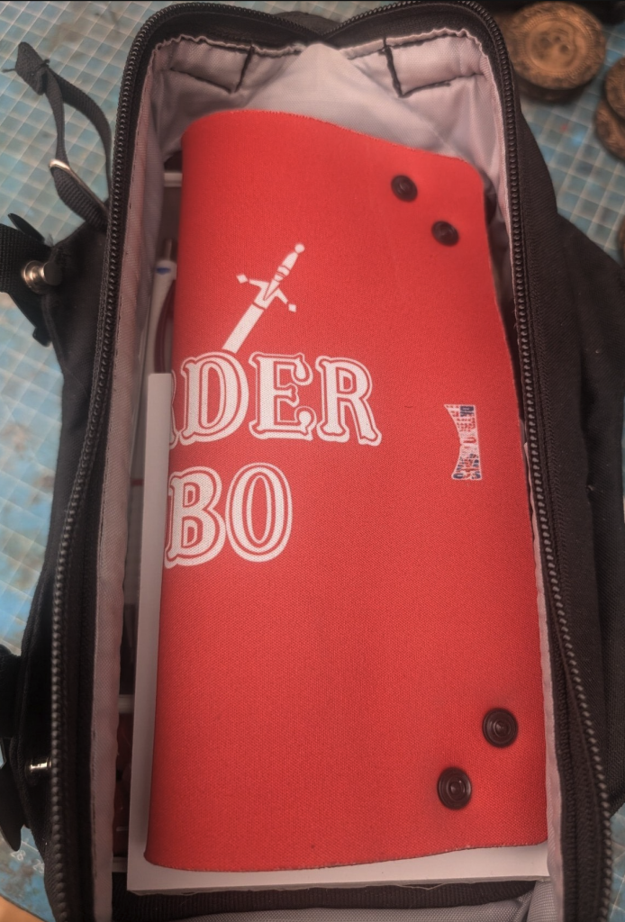 The bag is open, and there is a bright red neoprene mat that can be formed into a dice tray. It is has the words "MURDER HOBO" written on it, although one can only see "DER BO", as the mat is folded in half