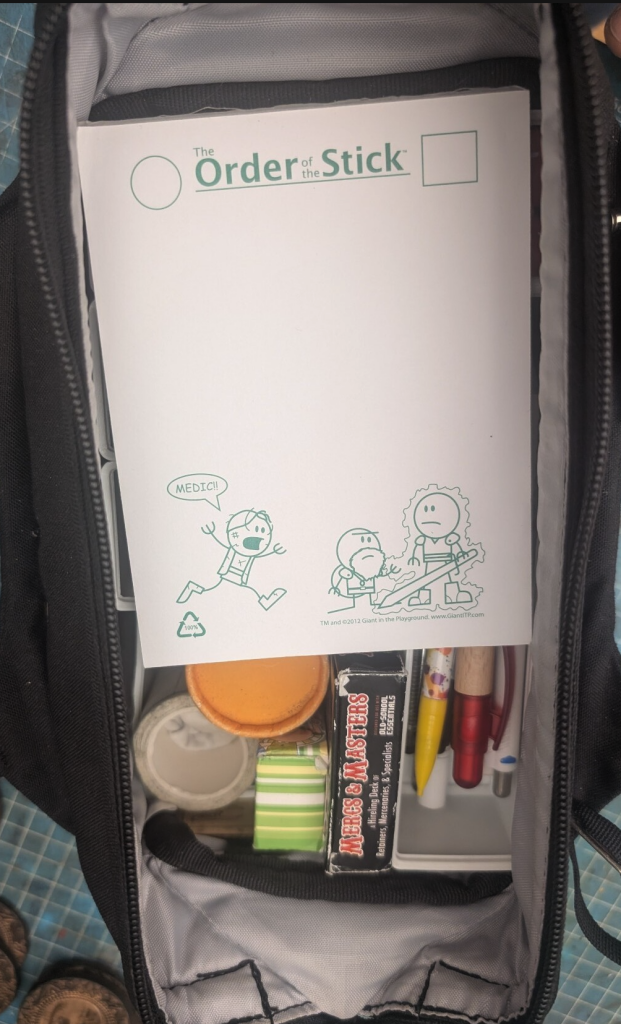The neoprene mat has been taken away, now one sees a few pens, rolls and a notepad from "The Order of the Stick", with Elan the bard  apparently banged up, running to the dwarf shouting for a "MEDIC!!"