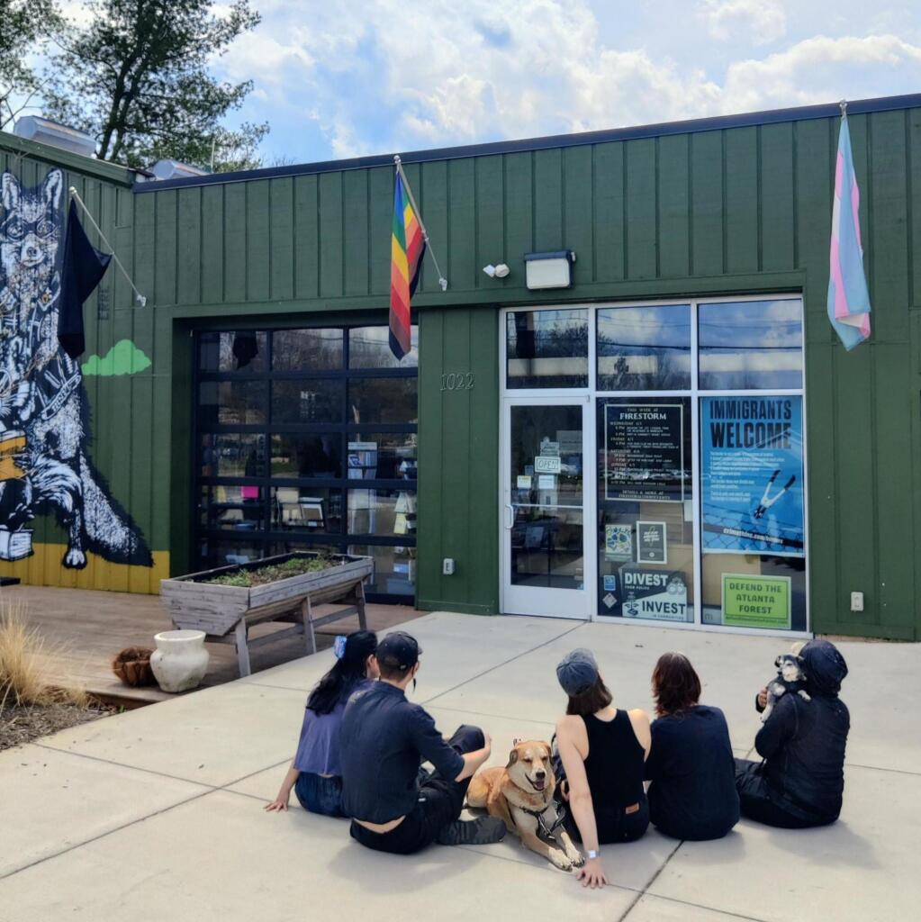 A collective photo taken at Firestorm Books. Our team, including five humans and two dogs, is sitting on the sidewalk in front of the building looking relaxed. The sky is bright but cloudy and flags on the building hang without any indication of wind. The building is painted forest green and various posters and signs are visible in the windows, which reflect the sky. The largest reads "Immigrants Welcome."