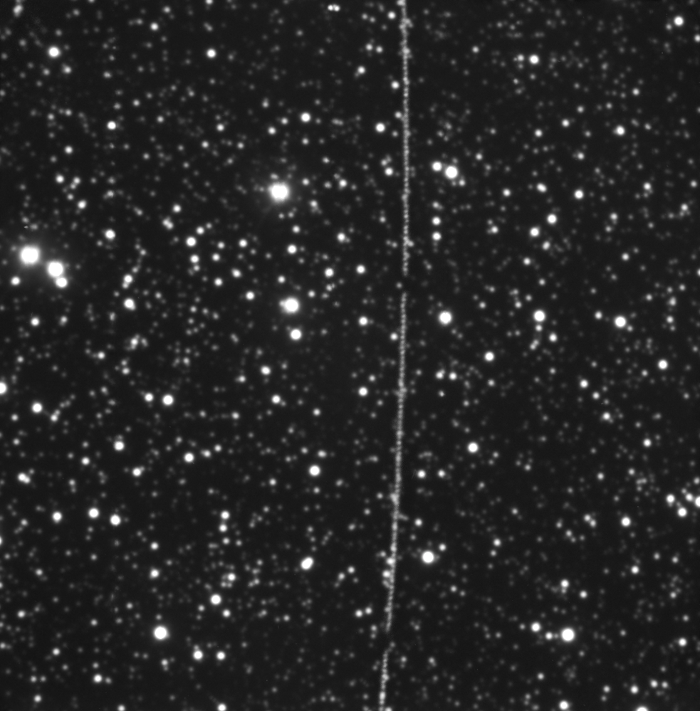 Black and white telescope image of a star-filled sky, with a bright vertical streak cutting through the centre, left by the passage of the Artemis II Orion module