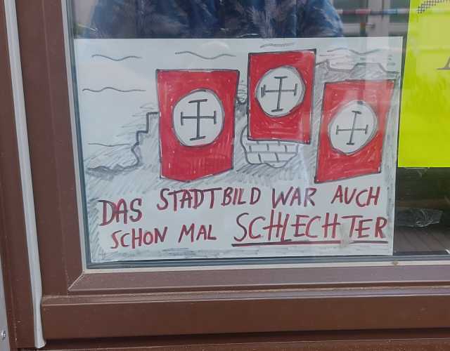 Im Murx ausgehängtes gezeichnetes Bild zum Thema Stadtbild. Ruinen Rot, Schwarz, Weise Banner. Dazu die Rote Aufschrift: Das Stadtbild war auch schon mal schlechter