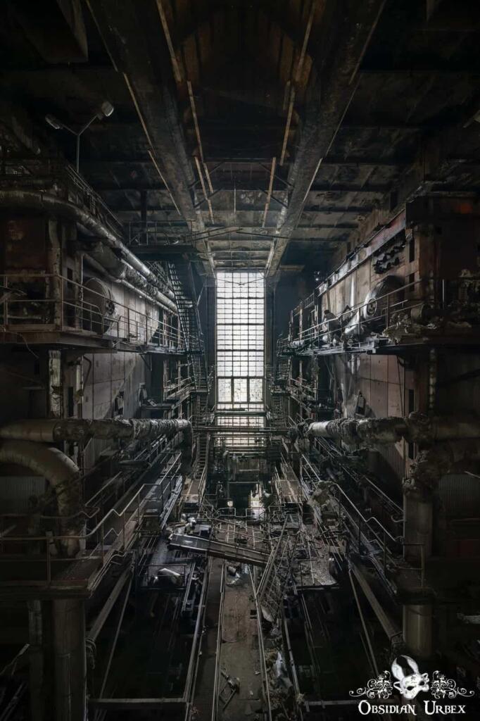 An atmospheric photo captures a vast, abandoned industrial power plant interior with multiple levels of derelict walkways and pipes leading towards a large, distant window.