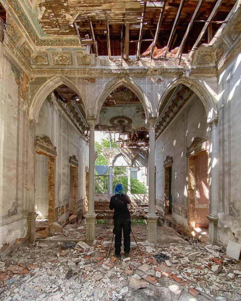 A person in a blue hat photographs the interior of a majestic, ruined palace in Lebanon, its ornate ceiling and arches crumbling amidst scattered debris.