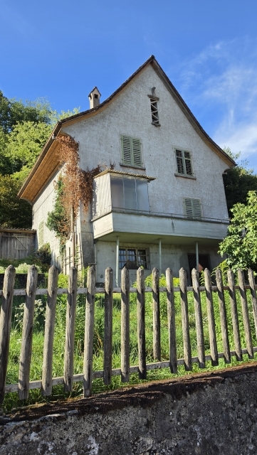 Ein helles Haus mit Satteldach, grünen Fensterläden und Balkon steht hinter einem Holzzaun.