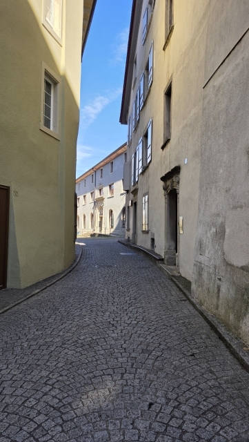 Eine schmale gepflasterte Gasse verläuft zwischen mehrstöckigen hellen Gebäuden unter blauem Himmel.
