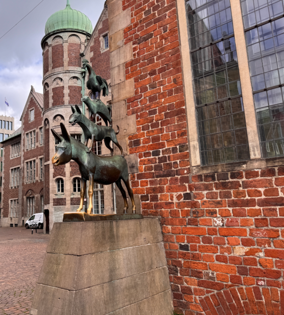Im Vordergrund steht eine bronzene Skulptur, die vier Tiere übereinander zeigt: Ein Esel bildet die Basis, darauf stehen ein Hund, eine Katze und ein Hahn. Die Skulptur befindet sich auf einem steinernen Sockel vor einer roten Backsteinmauer mit großen Fenstern. Links im Hintergrund ist ein historisches Gebäude mit einem runden Turm und grünem Kuppeldach zu sehen. Der Platz ist gepflastert und wirkt ruhig.