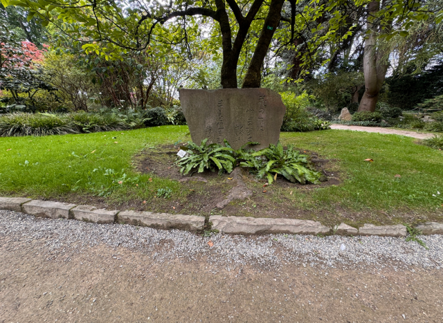 Im Vordergrund befindet sich ein rechteckiger, steinerner Gedenkstein mit eingraviertem Text, der auf einer kleinen Rasenfläche unter einem ausladenden Baum steht. Der Stein ist von Farnpflanzen umgeben. Im Hintergrund sind weitere Bäume und Sträucher zu sehen. Der Boden vor dem Stein besteht aus Kies und einer niedrigen Steinbegrenzung.