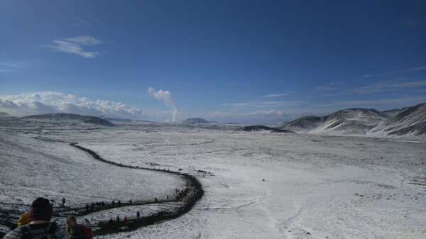 A bit of snow-covered flat land, surrounded by hills. A trail winding from the left all the way to the photographer, plenty of people on it. Far away, a small, thin mushroom cloud.