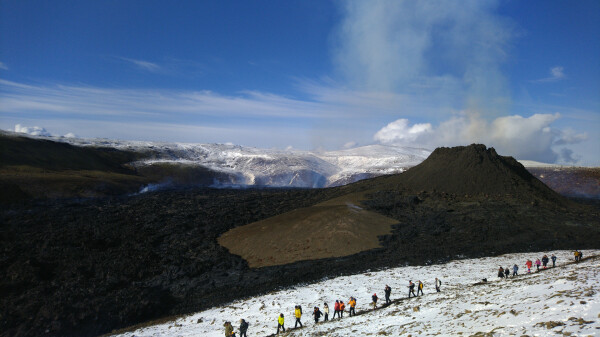 Volcano cone spewing hot fumes, with a black solidified lava field surrounding a small mound. The mound has cracked somewhat and there is a bit of gas visible escaping.

A stream of hikers visible beside it.