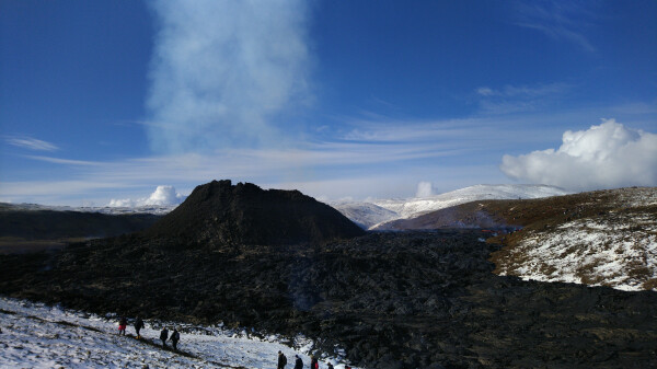 Another part of the lava field visible. Older and cooler. People coming up close to it, snow right next to it and a bit on it.

Volcano cone in the background.