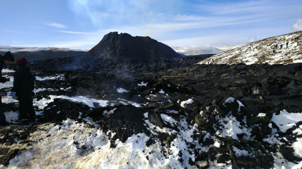 Close-up of the cool lava field, with some snow on it. People up very close to it. Volcano cone in the background.