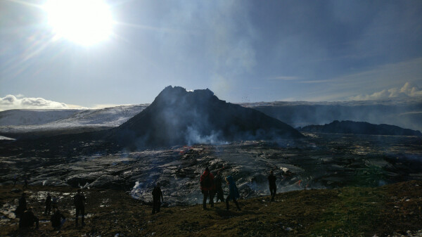 Black volcano cone, steaming, in the middle of a large lava field, in sharp sunlight under a blue sky. People here and there, looking at the volcano.