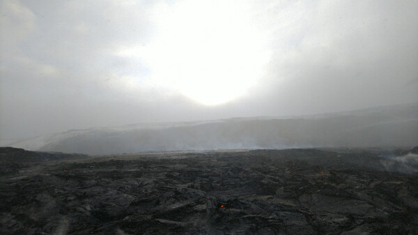 Heavy grey sky, with sun somewhat shining through the clouds. Lava field, black but steaming, a few embers visible here and there. Foggy hills in the background.