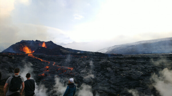 Volcanic eruption (two individual vents) in the background to the left. Lava flow over a cooled-down lava field, a lot of steaming and fuming all around. A few people watching the volcanic show. 