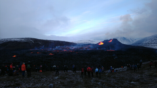 Erupting volcano in the background, behind it some snowy hills.

Large black lava field in the middle, with a lot of red and orange embers all over it.

About two dozen people watching the spectacle.