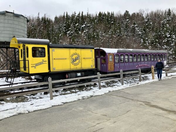 A train composed of a short yellow engine reading “Agiocochook” and “M4” in the back, and a purple wooden coach car in the front. The train is on a steep track with a wooden water tower and snowy conifers in the background.