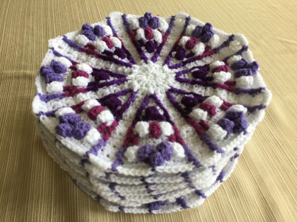 A pile of 9 octagonal crochet pieces. They have a triangular pattern on each side with puffs between two lines. The pattern has 4 different shades of purple radiating from the center on a white background.