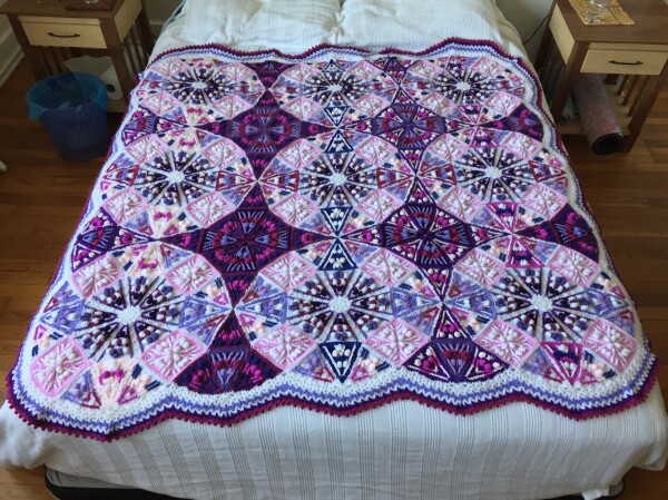 A crochet blanket spread out on a bed. There are 9 circles of pieces made of light colors with pieces made of dark colors between, with a striped border all around. The overall impression is pink and purple.