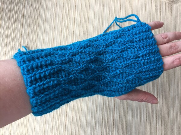 My left hand wearing a turquoise fingerless glove with an alternating ripple pattern along the length. There are strings hanging off where I haven’t sewn in the ends.