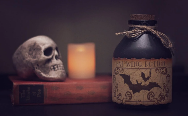 In the foreground on the right, a black matte potion bottle with a cork stopper and twine tied around the neck. The label advertises 'Bat Wing Potion' and has an illustration of bats and swirls against spiderwebs. Out of focus in the background, a small human skull and a glowing candle rest on top of an old book.