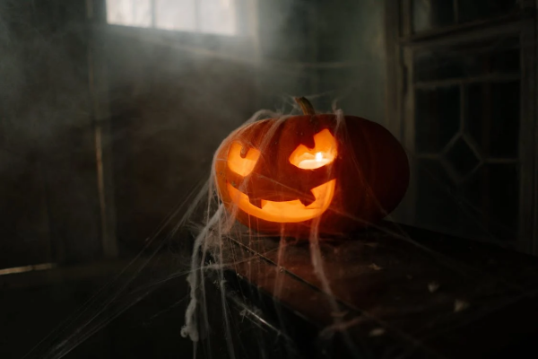 Spooky photograph of a jack-o-lantern lit with a candle and draped with cobwebs resting on an old chest. The room behind is dark, and dim light coming from a high window lights up the mist in the room.