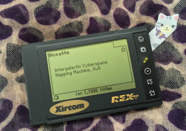 a pcmcia organizer with a little kitty sticker peering from behind it. the lcd reads 'discatte, intergalactic cyberspace napping machine, 0w0'