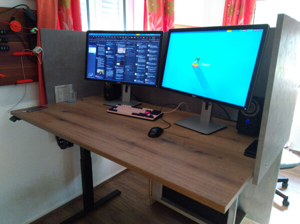 Top view of a wooden work desk, with two computer screens, a keyboard and a mouse on it. The desk has three wooden walls of ~60cm height on the left, right and back side, covered with gray concrete-like print.