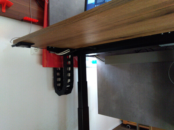 Bottom view of a height-adjustable desk with a ~6cm wide cable chain connecting the table to the wall.