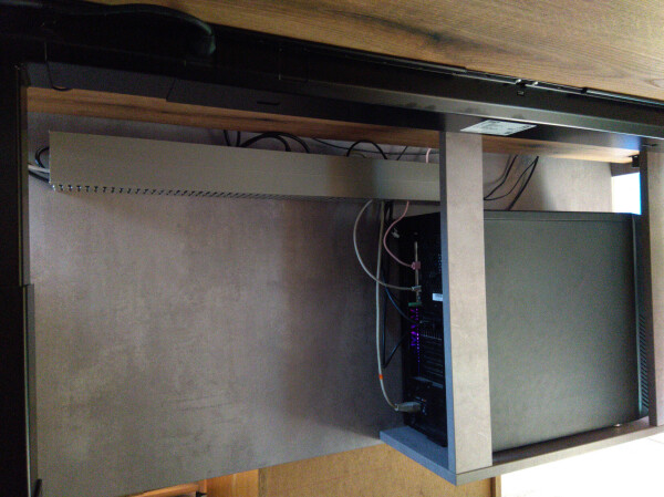 Bottom side of a work desk, with a wooden wall at the back side of the table that reaches around 60cm below the table, and mounted to this wall is a cable conduit and a mount for the desktop computer.