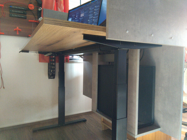 A height-adjustable desk, picture taken from below, where a mount for the desktop computer and a cable chain connected to the wall can be seen.