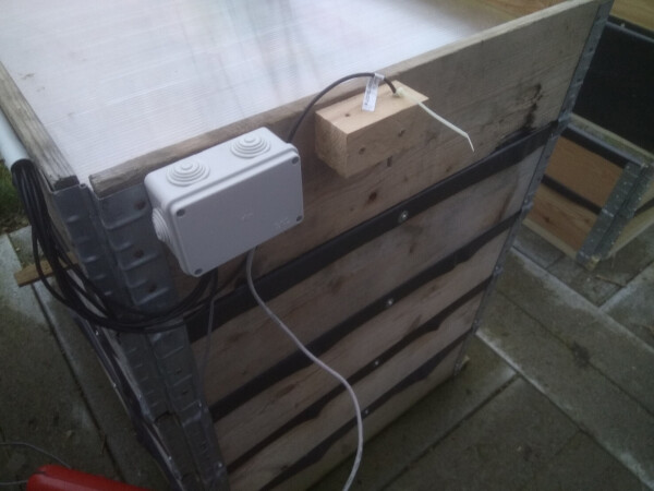 A raised bed with a water-proof plastic box mounted on the outside of its wooden frame, and some cables coming out of the plastic box.