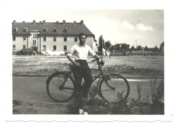 A man in white shirt and dark trousers standing by a bicycle. Behind him a meadow and a three-story building.