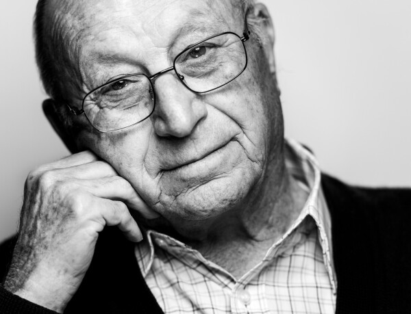 A portrait photo of an older man in a glasses. His head is leaned against his hand.