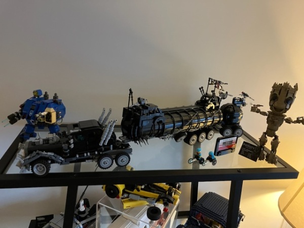 A top glass shelf with 3 LEGO sets displayed: a blue Warhammer 40,000 Ultramarines Dreadnought MOC, a black Mad Max’s War Rig MOC taking the whole length of the shelf, and a brown Guardians of the Galaxy baby Groot figure.