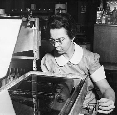 Katharine Burr Blodgett sat working at an unidentified machine with a large glass surface. She has dark hair in a bun, glasses and wears a light colour short sleeved dress with whit collar and cuffs at elbow, her left hand operating a handle on the machine. Black and white photo 