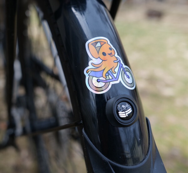 urbanists.video logo as a sticker placed on rear fender of bike. The logo is an orange, happy cuttlefish riding a purple bike.