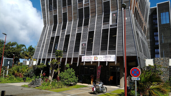 The library of Saint-Joseph, a contemporary building in appaerance made of wood with the shape of a geometrical hourglass patched with many windows. Green palm trees and tropical plants are all around it. In the front, the banner of the "Reunion des Communs"