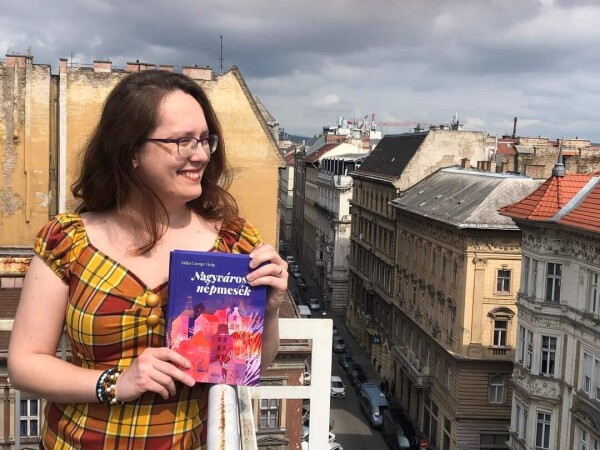 Photo of me (mid-30s, red hair, glasses, yellow checkered top) on a rooftop, holding the book (purple cover with pink and orange house motifs), with a Budapest street in the background. 