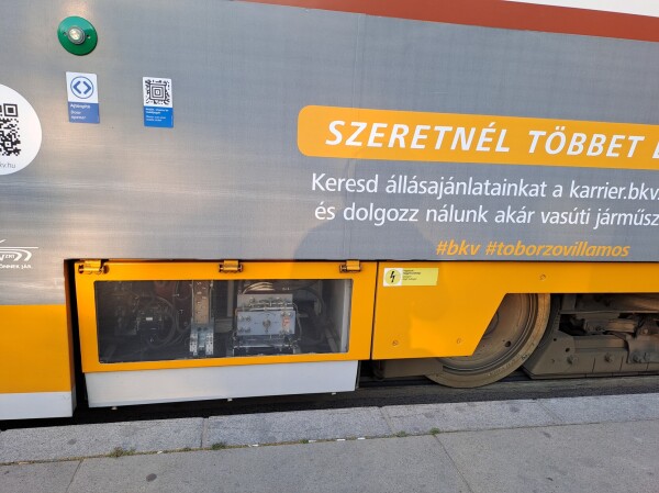 Clear panel by the wheels, showing ehat I assume is the braking mechanism? I am very bad at this. Partial sign says "would you like to know more? Look for our job advertisements and work for us" in Hungarian.