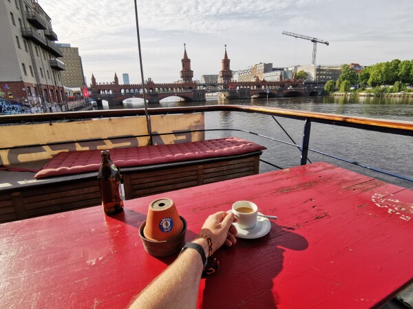 Ein Tisch auf dem Oberdeck eines Hotelschifs auf der Spree in Berlin. Auf dem Tisch steht eine Kaffeetasse, die Ralf in der Hand hält. Im Hintergrund ist die Spree und die Oberbaumbrücke zu sehen. 