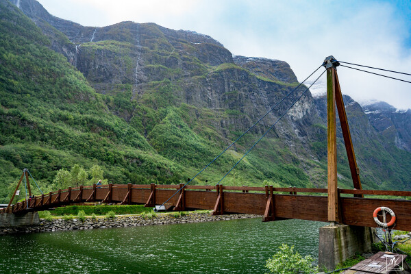 Color photo taken near one end of a thin wooden suspension bridge spanning across water that is in a narrow valley with tall, steep sides. Coming off the mountainsides high above are thin waterfall streams. The sky above is mostly cloudy but with some patches of blue. 