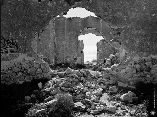 A black-and-white photo from inside the ruins of one of the buildings looking out through a door opening out to sea.