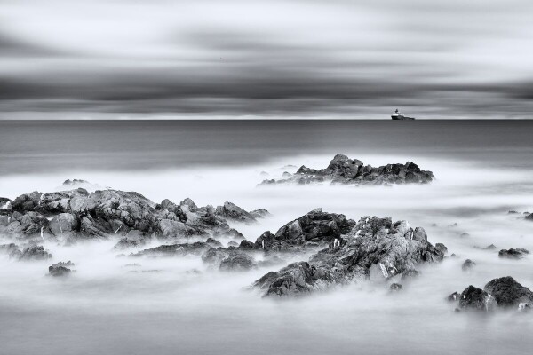 Long-exposure seascape off the rocks out to sea. There's a ship on the distant horizon. The rough waves have been smoothed into a mist. The sky has menacing clouds.