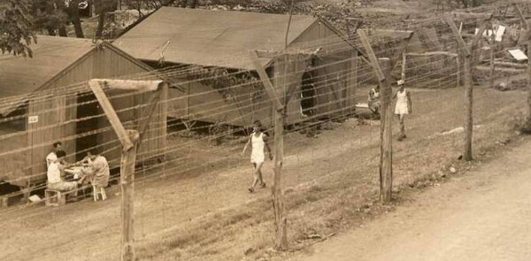 Photo of Japanese-American internees walking behind barbed wire fence that is well over the height of a person, past wooden barracks