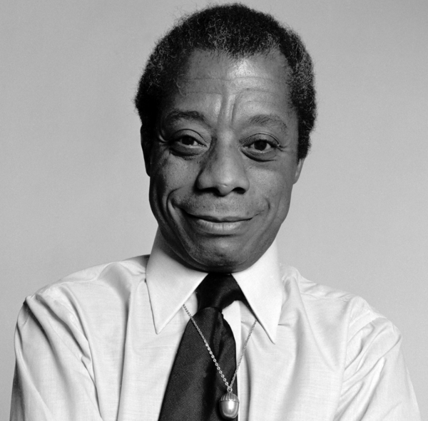 Black and white photo of a smiling man in white shirt and black tie; this is James Baldwin